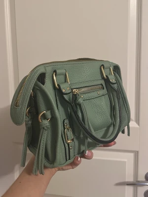 Grön handväska med gulddetaljer - Snygg grön handväska i skinnimitation med guldfärgade metalldetaljer och dragkedjor. Väskan har dubbla handtag, dekorativa spännen och tofsar på sidorna. Invändigt finns randigt foder och flera fack för smart organisering. Perfekt storlek för det viktigaste!