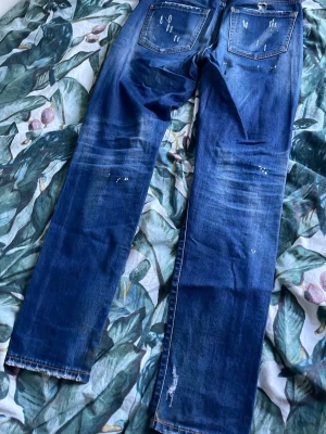 Blå distressed jeans från Dsquared2 - Säljer ett par blå jeans från Dsquared2 med straight fit och coola slitningar, hål och färgstänk för en edgy look. Jeansen har klassisk femficksmodell, normal passform och är gjorda i slitstarkt bomullsmaterial. Perfekta för dig som gillar streetwear och statement-plagg.