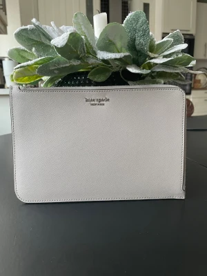 Grå kuvertväska från Kate Spade - Snygg grå kuvertväska från Kate Spade i skinn med stilren design och silverfärgad logga. Väskan har handledsrem, dragkedja och ett fack på baksidan. Insidan är ljus med hjärtmönster och har kortfack. Perfekt för essentials och enkel att bära med sig.
