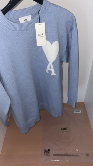 Ljusblå stickad tröja från Ami - Säljer en ljusblå stickad tröja från Ami Alexandre Mattiussi med ett vitt hjärta och bokstaven A på bröstet. Tröjan har rund hals, ribbade muddar och lång ärm. Perfekt för dig som vill ha en clean men ändå unik look.