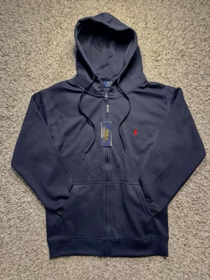 Ralph Lauren Zip Hoodie - Klassisk Marinblå Hoodie från Ralph Lauren. Gjord av mjukt och högkvalitativt material med en smidig kedja. Är i storlek M, men den sitter som S. Nypris ligger på 1699