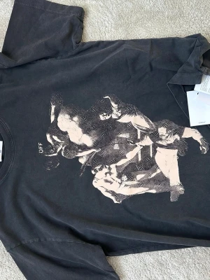 Svart Off-White t-shirt med konsttryck - Svart t-shirt från Off-White i bomull med stort grafiskt konsttryck framtill och tryckt logga på ryggen. Klassisk passform med rund hals och korta ärmar. Snygg streetwear-vibe och coola detaljer som gör den unik.