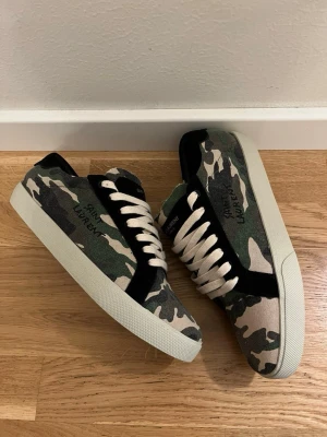 Saint Laurent sneakers i camo - Snygga Saint Laurent sneakers med camouflage-mönster i grönt, beige och svart. Skorna har vita snören, vit sula och detaljer i svart mocka. Märkeslogga på sidan och tungan. Perfekta för dig som vill sticka ut med en cool streetstyle. Passar även storlek 45!