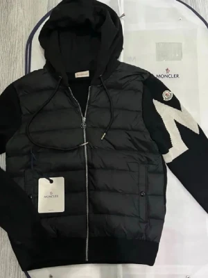 Svart pufferjacka från Moncler - Säljer en svart pufferjacka från Moncler med huva och dragkedja framtill. Jackan har stickade ärmar med vit zigzag-detalj och Moncler-logga på ena ärmen. Kroppen är quiltad och vadderad, medan ärmarna är i mjukt stickat material. Snygg och sportig look.