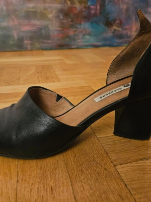 Svarta pumps från Blankens i skinn - Stilrena svarta pumps från Blankens i äkta skinn med spetsig tå och bred klack. Skorna har öppen insida och unik detalj baktill på hälen. Perfekta för dig som vill ha en klassisk men ändå edgy look.