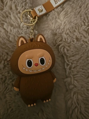 Brun plånbok i form av söt figur - Säljer en liten brun plånbok i form av en gullig figur med öron och stort leende. Plånboken har dragkedja på sidan och nyckelring med rem, perfekt att hänga på väskan eller använda som nyckelring. Materialet är mjuk syntet och designen är riktigt lekfull.
