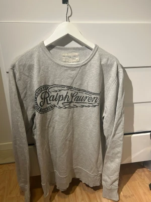 Grå sweatshirt från Ralph Lauren - Snygg grå sweatshirt från Ralph Lauren Denim & Supply med stort tryck på bröstet. Tröjan har rund hals, långa ärmar och ribbade muddar vid ärmslut och nederkant. Perfekt för en chill och avslappnad stil.