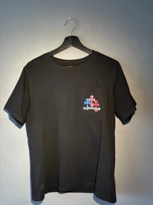Svart Chrome Hearts t-shirt med färgglada kors - Svart t-shirt från Chrome Hearts med färgglada kors i blått, rött, grönt, gult, rosa och vitt på ryggen och ett mindre tryck på bröstfickan. Klassisk passform och rund halsringning. Perfekt för dig som gillar streetwear och unika prints.