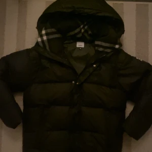 Svart pufferjacka från Burberry - Säljer en svart pufferjacka från Burberry med klassiskt rutigt foder i huvan. Jackan har en stor huva, dragkedja och tryckknappar framtill samt Burberry-logga på ärmen. Perfekt för kalla dagar och riktigt snygg till streetwear-stil.
