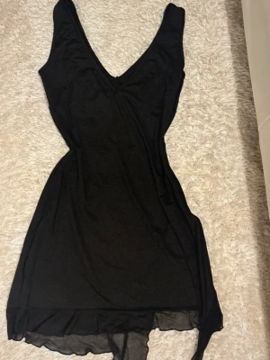 Svart klänning med v-ringning - Svart klänning med djup v-ringning och asymmetrisk nederkant i tunt, skirt material. Klänningen är ärmlös och har en figurnära passform som framhäver midjan. Perfekt för dig som gillar stilrena och enkla plagg med lite edge.