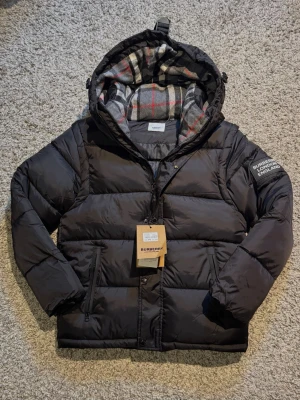 Svart Burberry Jacka - Svart pufferjacka från Burberry med huva och klassiskt rutigt foder i grått, vitt och rött. Jackan har dragkedja, knappar med Burberry-logga och en patch på ärmen med texten 'Burberry London England'. Perfekt för kalla dagar. Ärmarna kan tas bort och jackan kan göra till en väst.