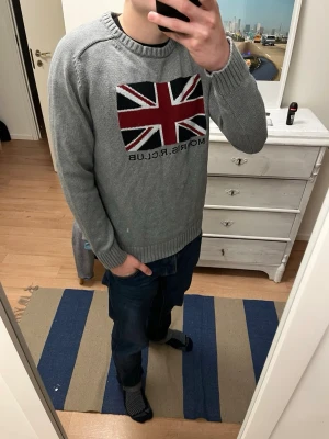 Grå stickad tröja Morris R Club - Säljer en grå stickad tröja från Morris R Club i storlek L. Tröjan har ett stort Union Jack-motiv i rött, svart och vitt på bröstet samt texten 'Morris R Club' under. Rund hals och ribbade muddar vid ärmslut och nederkant. Perfekt för höst och vinter. L men passar även M
