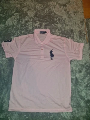 Sportpiké från Polo Ralph Lauren M - Säljer en rosa pikétröja från Polo Ralph Lauren i storlek M. Klassisk krage, knäppning med två knappar och stor mörkblå logga broderad på bröstet. Pikén har korta ärmar och är tillverkad i polyester