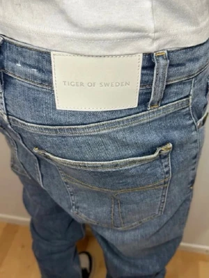 Blå jeans från Tiger of Sweden - Säljer ett par klassiska blå jeans från Tiger of Sweden. Jeansen har normal passform, raka ben och fem fickor. Snygga kontrastsömmar och en stor vit patch med logga bak i midjan. Tillverkade i slitstarkt denimtyg som ger en tidlös look. Hör av er vid minsta lilla fundering ! 