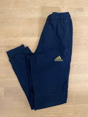 Mörkblå Real Madrid Adidas mjukisbyxor - Säljer ett par mörkblå mjukisbyxor från Adidas med Real Madrid-logga i guld på ena benet och Adidas-logga i guld på andra benet. Byxorna har resår i midjan och vid bensluten samt två sidofickor. Perfekta för chill eller träning.