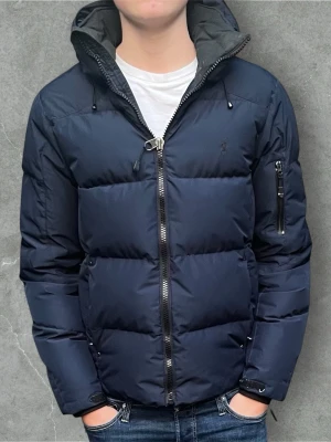 Mörkblå pufferjacka från Polo Ralph Lauren - Säljer en mörkblå pufferjacka från Polo Ralph Lauren i storlek XS. Jackan har hög krage, huva och grov dragkedja framtill. Fylld med dun och har en clean, oversized look som är perfekt för kalla dagar. Snygg och enkel design med diskret logga på bröstet. Skicket är bra med ytterst lite skavanker. Bild kan fås om det önskas! Köpt i Ralph Lauren butik.