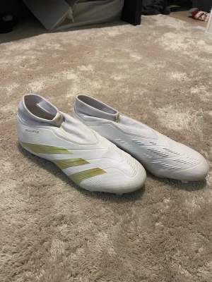 Adidas Predator vita fotbollsskor - Tvättar skorna innan köp. Säljer ett par vita Adidas Predator fotbollsskor med gulddetaljer och strumpliknande skaft. Skorna har en modern, strömlinjeformad design utan snörning och är tillverkade i syntetmaterial för bästa bollkontroll. Perfekta för dig som vill sticka ut på planen. Skickas endast med PostNord. Om annat fraktsätt väljs av Plick, avbryts köpet.