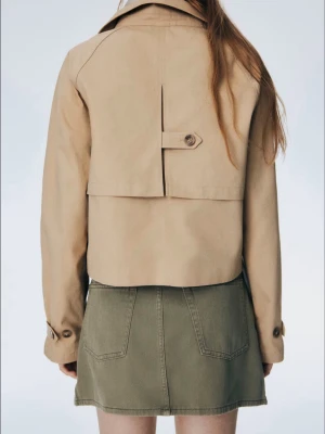 Beige kort trenchcoat med knappar - Snygg beige trenchcoat från H&M med dubbelknäppning och klassisk krage. Jackan är croppad och har långa ärmar med dekorativa detaljer vid mudden. Perfekt för dig som vill ha en stilren och modern look. Knappt använd! Tar emot byten