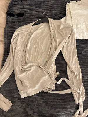 Beige omlottkofta med knytband - Säljer en beige omlottkofta med långa ärmar och knytband i midjan. Koftan är tunn och mjuk, perfekt att slänga över en topp för en chill och snygg look. Materialet känns lätt och skönt mot huden.