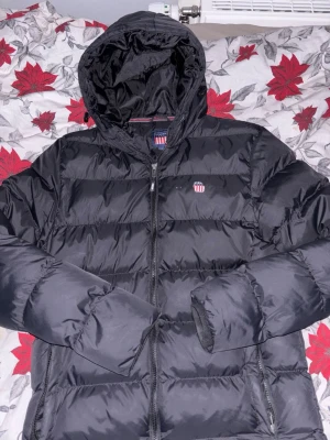 Svart pufferjacka från Vinson - Svart pufferjacka från Vinson med huva och dragkedja framtill. Jackan har ett broderat Vinson-emblem på bröstet och är quiltad för extra värme. Perfekt för kalla dagar och har en clean, sportig look med diskreta detaljer.