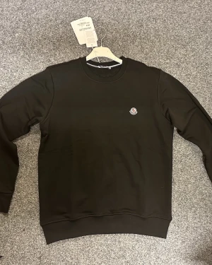 Svart sweatshirt från Moncler - Säljer en svart sweatshirt från Moncler med klassisk rund hals och Moncler-logga broderad på bröstet. Tröjan har långa ärmar och ribbade muddar vid ärmslut och nederkant. Tillverkad i mjuk bomull för skön känsla.
