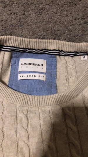 Beige kabelstickad tröja Lindbergh M - Snygg beige kabelstickad tröja från Lindbergh i relaxed fit. Tröjan har rund hals, ribbade muddar och ett klassiskt kabelmönster över hela plagget. Perfekt för dig som gillar stilrena och tidlösa plagg med lite extra struktur.