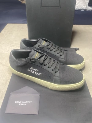 Svarta Saint Laurent sneakers - Snygga svarta sneakers från Saint Laurent med handskriven logga på sidan. Skorna har ovandel i canvas och detaljer i skinn, samt en vit platt gummisula. Klassisk låg modell med snörning och diskreta Saint Laurent-detaljer på plös och sula.