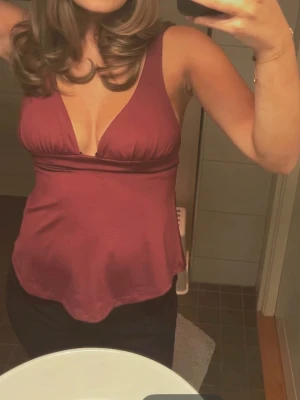 Vinröd v-ringad topp med knytning - Säljer en vinröd topp med djup v-ringning och breda axelband. Toppen har omlott-detalj och knyt i ryggen för extra snygg passform. Materialet är mjukt och har ett lätt glansigt utseende, perfekt för dig som gillar stilrena och trendiga plagg.