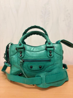 Grön handväska i skinn - Cool grön handväska  i mjukt skinn med flätade handtag, justerbar axelrem och snygga metalldetaljer. Väskan har dragkedja upptill och framtill samt dekorativa nitar. Perfekt storlek för det viktigaste och ger en edgy vibe till din outfit.