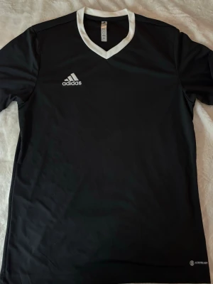 Svart Adidas  t-shirt - Svart t-shirt från Adidas med vit v-ringad krage och broderad logga på bröstet. Tillverkad i lätt och funktionell polyester som andas, perfekt för sport och träning.