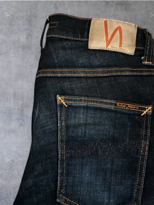 Mörkblå jeans från Nudie Jeans, W28 L32 - Snygga mörkblå jeans från Nudie Jeans med klassisk femficksdesign och kontrastsömmar i orange. Jeansen har en rak passform och är tillverkade i slitstarkt denimtyg av bomull. Perfekta för dig som gillar stilrena och tidlösa jeans med schyssta detaljer.