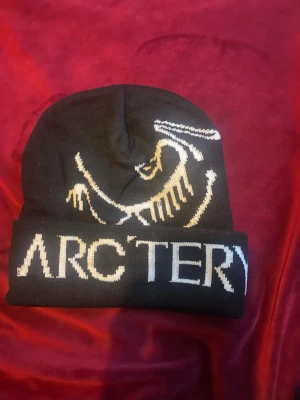 Svart Arc'teryx mössa med logga - Snygg svart mössa från Arc'teryx med stor vit logga och text framtill. Stickad i mjukt material som håller dig varm och ger en clean look. Perfekt för dig som gillar outdoor och streetwear. Passar både tjejer och killar.