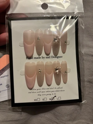 Handgjorda lösnaglar med dekoration - Säljer ett set handgjorda lösnaglar i nude och ljusrosa toner. Naglarna har både matt och glittrig finish, vissa dekorerade med små stenar och en 3D-blomma. Formen är almond och långa. Setet inkluderar nagelfil, applikationsverktyg och klisterkuddar.