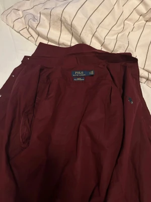 Vinröd skjorta från Polo Ralph Lauren - Säljer en vinröd skjorta från Polo Ralph Lauren i 100% bomull. Skjortan har klassisk krage, knäppning framtill med vita knappar och den ikoniska broderade loggan på bröstet. Perfekt för dig som vill ha en stilren och tidlös look.