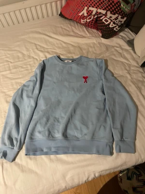 Ljusblå sweatshirt från Ami Paris - Säljer en ljusblå sweatshirt från Ami Paris med röd broderad logga på bröstet. Tröjan har rund hals, långa ärmar och ribbade muddar. Perfekt för dig som gillar stilren design med en twist.