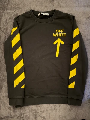 Off-White svart sweatshirt med gula ränder - Svart sweatshirt från Off-White med feta gula tryck på både fram- och baksida. Ärmarna har tydliga diagonala ränder och framtill finns Off-White-loggan med pil. Tröjan har rund hals och är långärmad, perfekt för dig som gillar streetwear och vill sticka ut. Storlek M