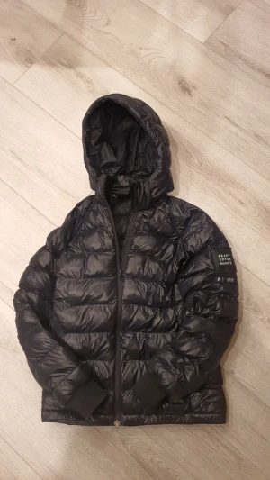 Svart dunjacka Peak Performance - Svart dunjacka från Peak Performance med huva och dragkedja. Jackan är quiltad och har ribbade muddar vid ärmsluten. Märkespatch på ena ärmen. Perfekt för kyliga dagar och har en clean, sportig look. Den har även ett litet hål som är övertäckt. Jackan har funkat bra för mig i alla väder. Den passar alla mellan ca 155-165cm.