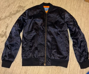 Mörkblå bomberjacka från Adidas - Snygg svart bomberjacka från Adidas med orange foder och klassisk ribbad krage. Jackan har dragkedja framtill, två fickor och diskret Adidas-logga på bröstet. Materialet är glansigt och känns lätt, perfekt för dig som vill ha en stilren och skön lool. 