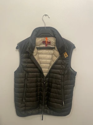 Olivgrön dunväst från Parajumpers - Snygg olivgrön dunväst från Parajumpers med quiltad design och hög krage. Västen har två dragkedjeförsedda fickor, gul detalj vid halsen och PJS-logga på ryggen. Insidan är ljusbeige och materialet är lätt och smidigt, perfekt för lager-på-lager under kyliga dagar.