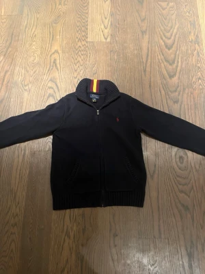 Mörkblå kofta från Polo Ralph Lauren - Mörkblå stickad kofta från Polo Ralph Lauren med dragkedja och ribbad krage med röda och gula ränder. Klassisk röd logga på bröstet och två fickor framtill. Perfekt för dig som gillar stilrena plagg med snygga detaljer.