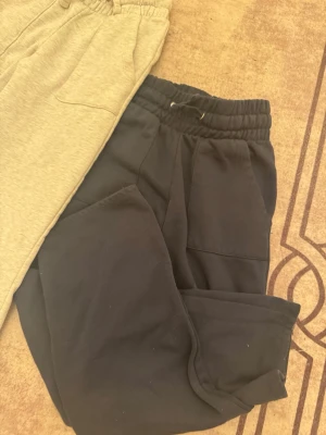 Gråa och svarta mjukisbyxor med snörning - Två par mjukisbyxor, ett par i ljusgrått och ett par i svart. Båda har elastisk midja med snörning och stora fickor framtill. Perfekta för chill dagar eller när du vill vara extra bekväm. Materialet är mjukt och känns skönt mot huden. Samt så är längden på båda 100cm