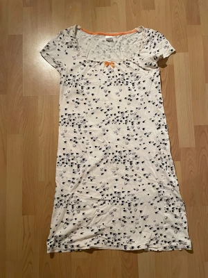 Vit långklänning med fågelmönster - Klänningen har korta ärmar, svart och vit fågelmönster över hela tyget och en orange rosett vid halsringningen.