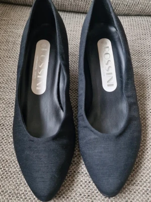 Svarta pumps från Rossini, storlek 39 - Stilrena svarta pumps från Rossini i textilmaterial. Skorna har spetsig tå och en klassisk, elegant form som passar till många olika outfits. Låg klack och mjuk finish. Tyget ligger inte helt limmat mot skon överallt (se bilder). Inget som jag tycker förstör utseendet men det är väl en smaksak. Inköptes innan livet kom emellan och jag fick inse att sneakers är smidigare 😂 Som ni ser på sulorna är de använda men endast vid ett par tillfällen. Tyget är lite skadat vid klackarna (se bild)