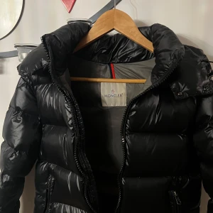Svart Moncler pufferjacka med huva - Svart pufferjacka från Moncler med glansig finish, huva och dragkedja framtill. Jackan har långa ärmar, två sidofickor med dragkedja och en cool Moncler-logga på ärmen. Insidan har ett unikt serietidningsmärke. Perfekt för kalla dagar. Pris kan diskureras vid snabbt köp!!!
