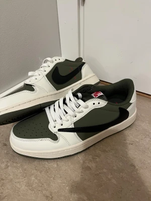 Nike Air Jordan 1 Low Travis Scott Olive - Nike Air Jordan 1 Low Travis Scott Olive med vit och olivgrön ovandel i skinn, svart omvänd swoosh och röda broderade detaljer på hälen. Perforerad tå, platt sula och snörning. Ikonisk Air Jordan-logga och Travis Scott smiley på baksidan. Extra skosnören medföljer.