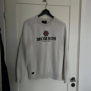 Vit stickad tröja från Morris - Vit stickad tröja från Morris med broderad logga och Union Jack-emblem på bröstet. Tröjan har rund hals, ribbade muddar och lång ärm. 