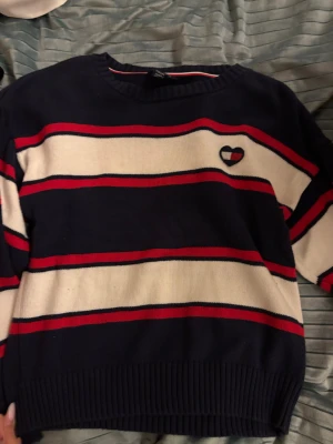 Randig stickad tröja Tommy Hilfiger - Snygg stickad tröja från Tommy Hilfiger med breda marinblå, vita och röda ränder. Tröjan har ett litet hjärtemblem på bröstet och ribbade muddar vid ärmslut och nederkant. Perfekt för dig som gillar klassisk stil med en twist.