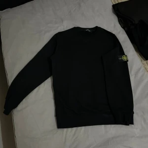 Svart sweatshirt från Stone Island - Svart långärmad sweatshirt från Stone Island med klassisk rund hals och ikonisk logopatch på vänster ärm. Tröjan har en clean look och är tillverkad i mjuk bomull som är skön att bära. Perfekt för dig som gillar streetwear och stilrena plagg.