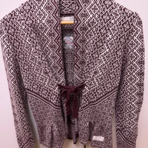 Mönstrad kofta från Odd Molly - Säljer en snygg kofta från Odd Molly i vinrött och vitt med stickat mönster. Koftan har knytband framtill, volangkant nertill och broderad logga på ryggen. Perfekt för dig som gillar unika detaljer och skön passform.