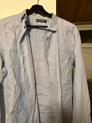 Ljusblå skjorta från Dolce & Gabbana - Säljer en stilren ljusblå skjorta från Dolce & Gabbana Gold. Skjortan har klassisk krage, långa ärmar och knappar framtill. Tillverkad i mjuk bomull som känns skön mot huden. Perfekt för dig som vill ha en clean och snygg look.